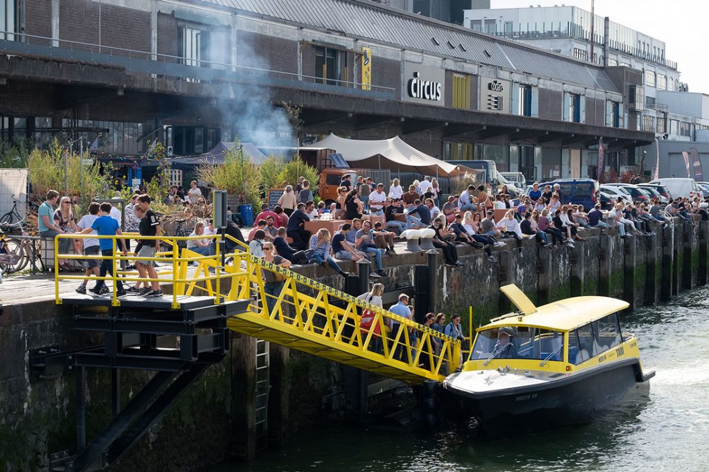 Mensen die op de kade zitten bij de fenix food factory op katendrecht in rotterdam tewrijl er een gele taxiboot aanmeert