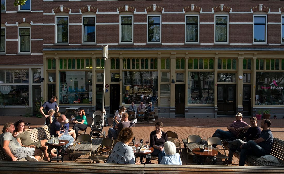 aanzicht van de delistraat aan het deliplein met ervoor mensen op bankjes en het terras op katendrecht na de renovatie van het deliplein